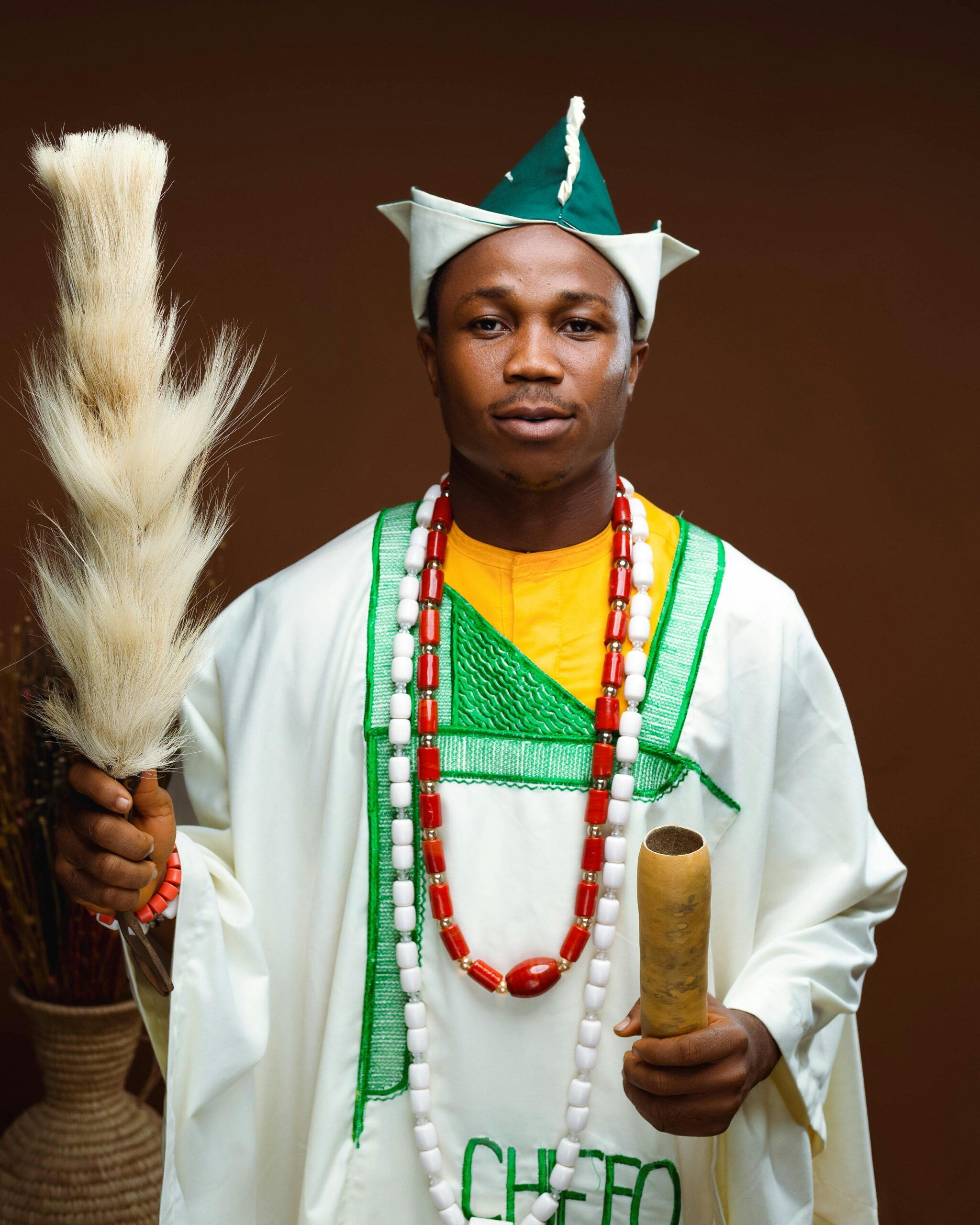African man in traditional Nigerian clothing holding cultural symbols in studio setting.